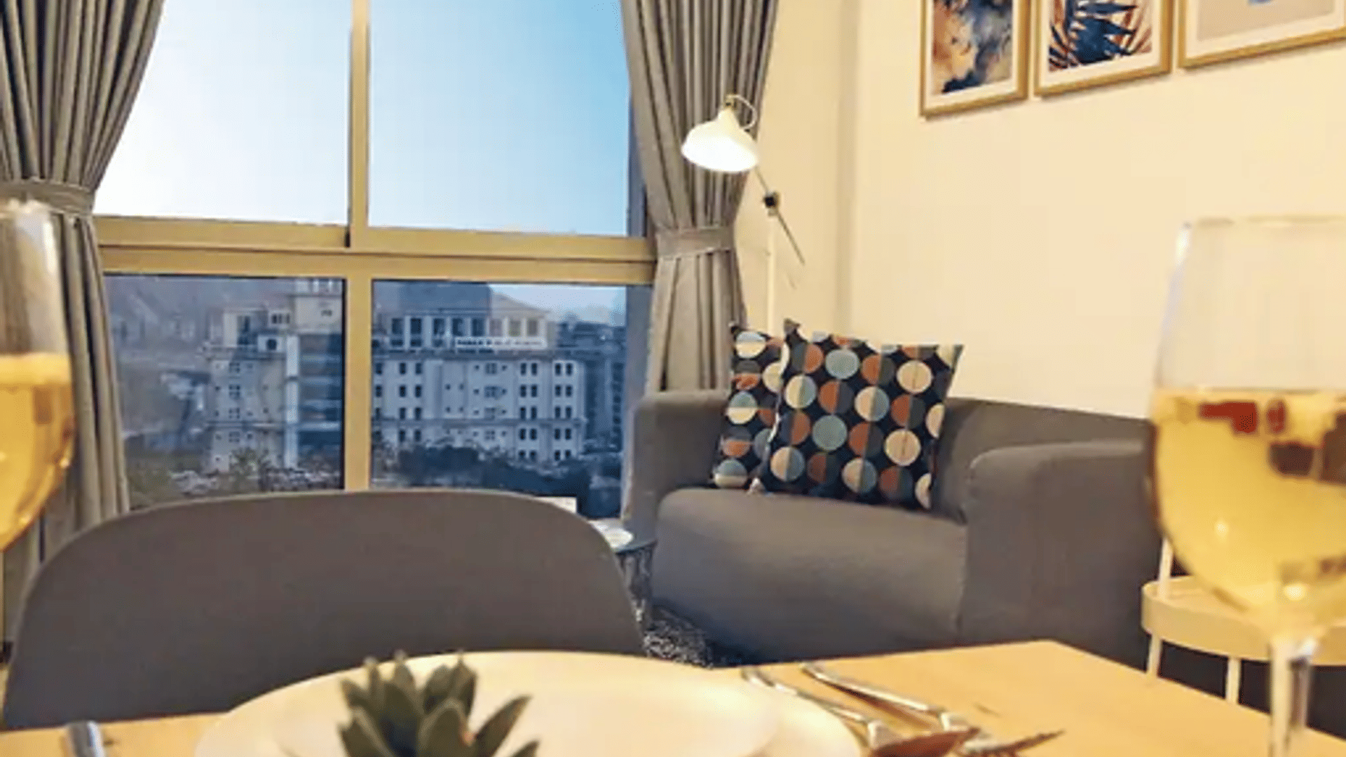A dining table set with plates, wine glasses, and a lit candle at Wabi-Sabi, Juhu, with a grey sofa and a large window overlooking the Mumbai skyline.