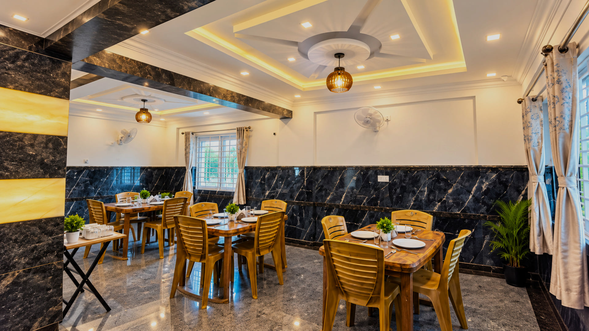 Dining area at Value Inn, Bangalore Airport with wooden tables set for a meal, black marble walls, pendant lamps, and a warmly lit tray ceiling