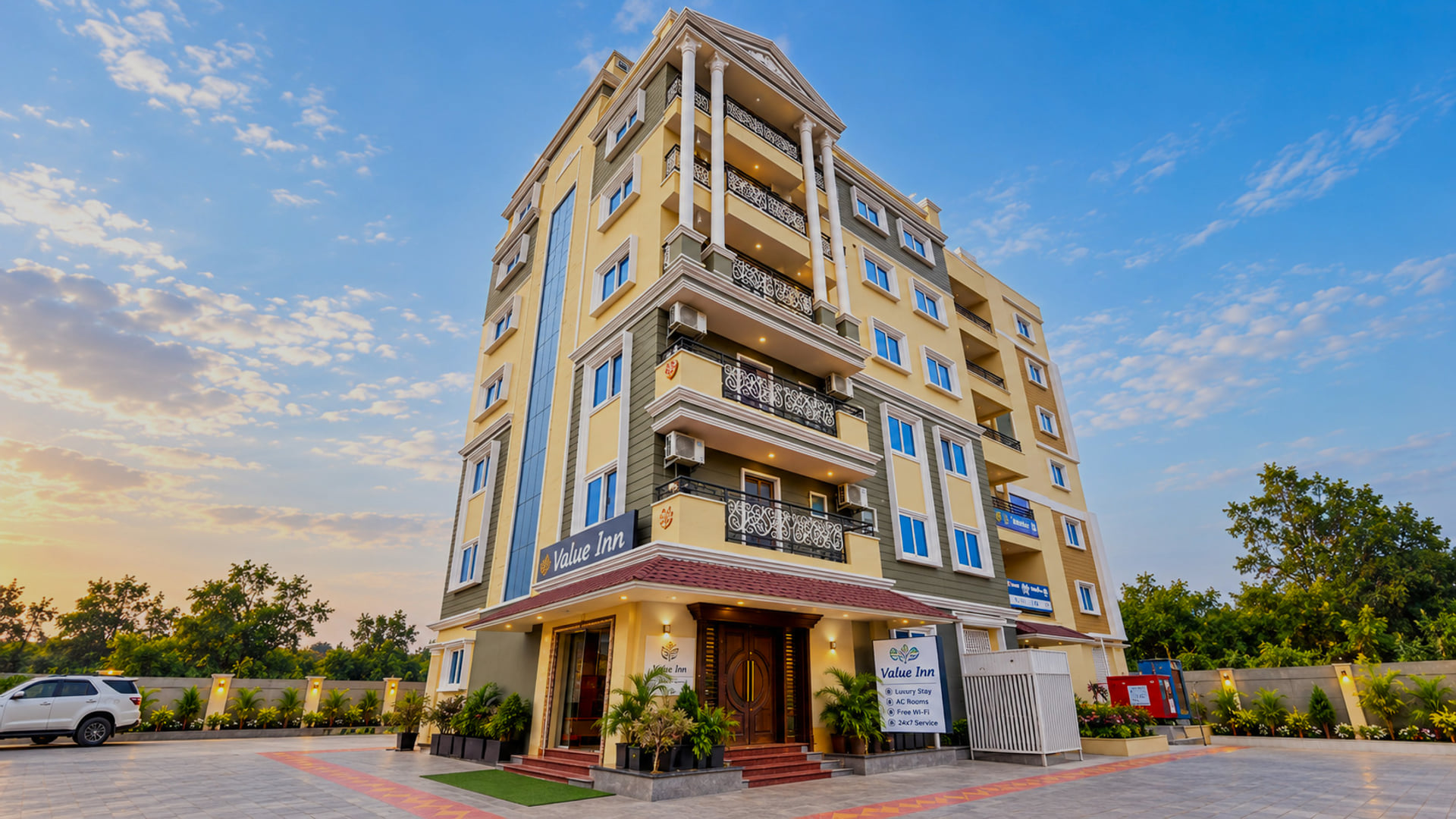 Front view of the Value Inn, Bangalore Airport building at sunset with its cream and yellow facade, ornate balconies, and plant lined entrance steps