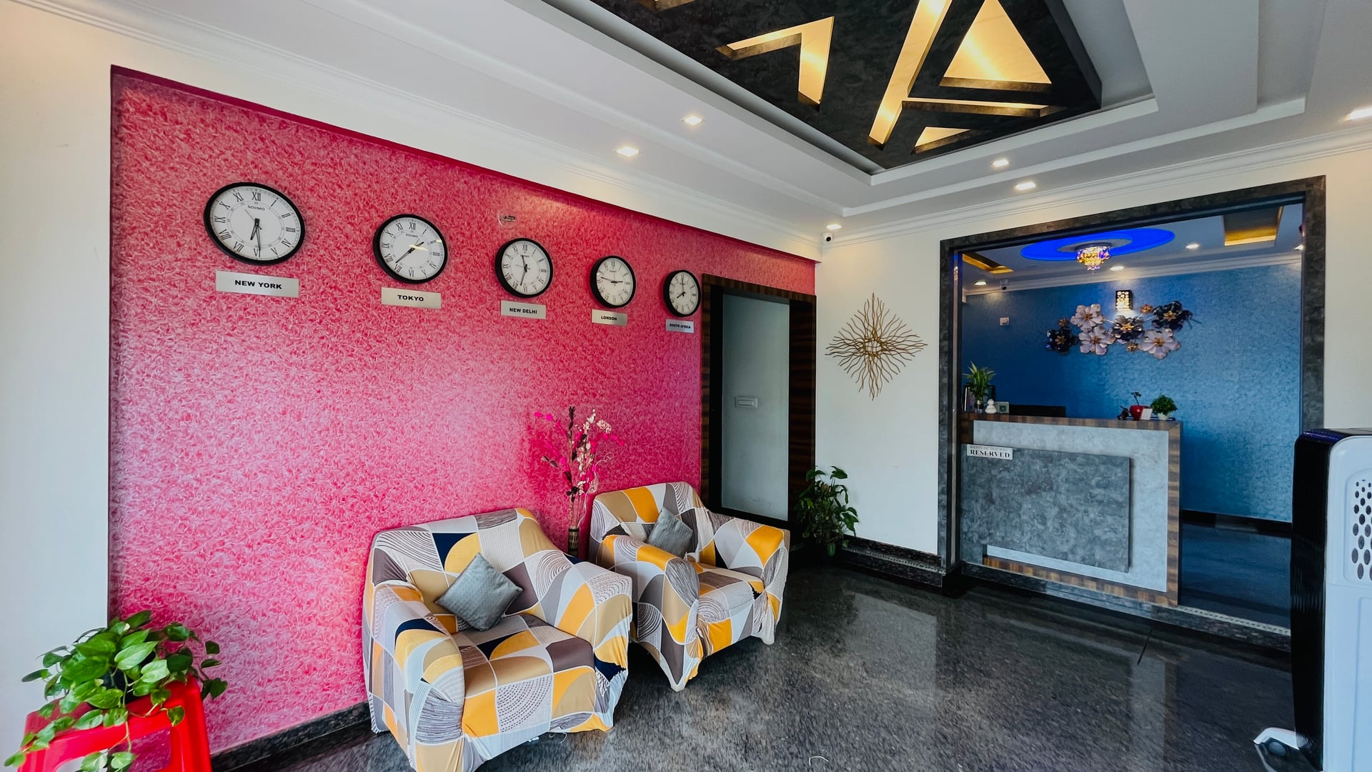 Lobby seating at Value Inn, Bangalore Airport with a patterned sofa, world clocks on a red feature wall, and the reception counter behind