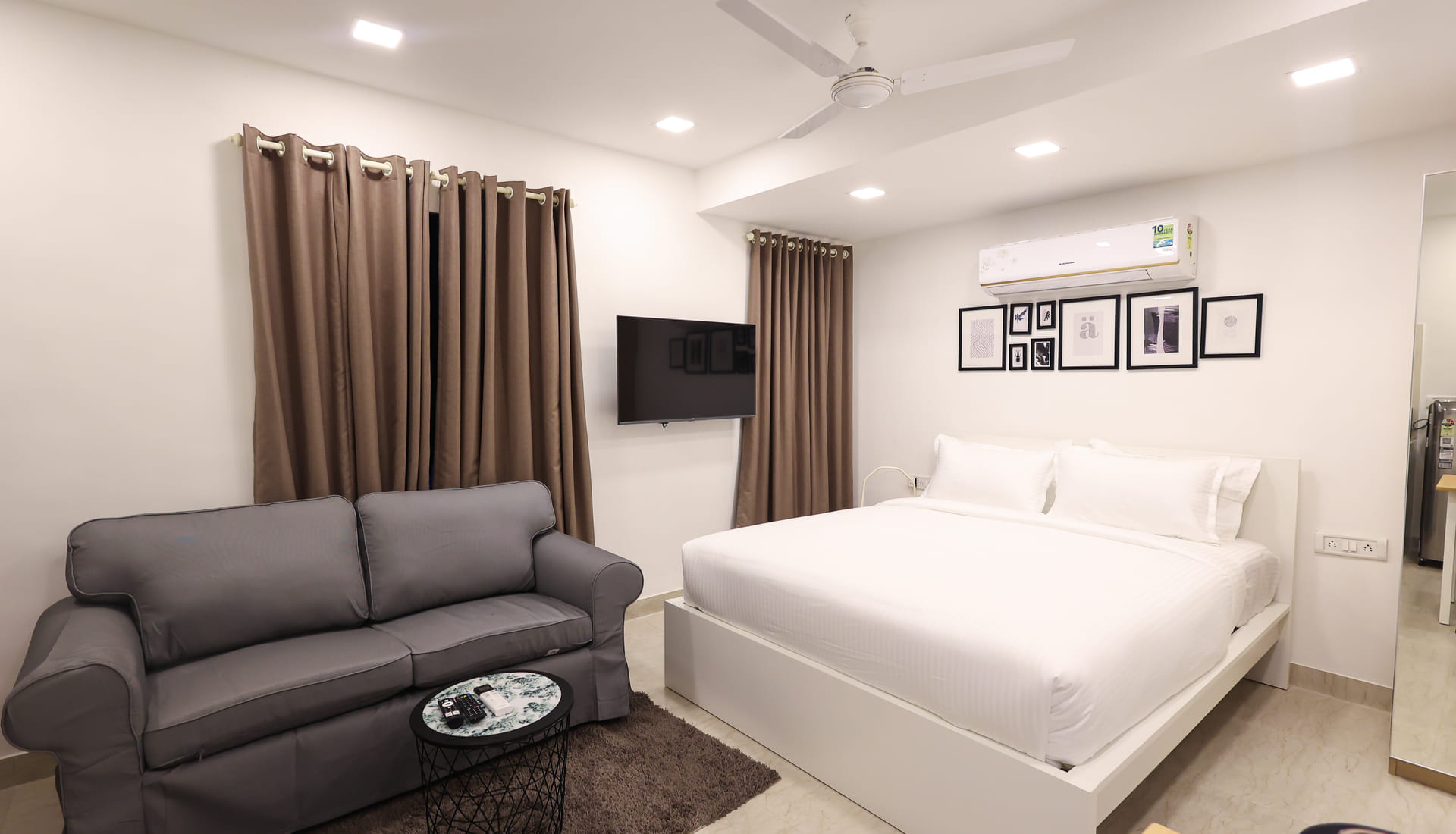 Studio at Wabi-Sabi, Juhu with a large bed made up in white linen, a grey two-seater sofa, wall-mounted TV, framed artwork and taupe curtains at two windows.