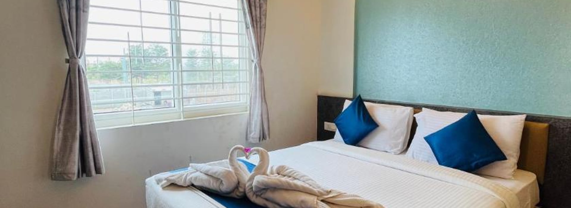 Queen bed with white linen, blue cushions, and a towel swan in the Standard Non-AC Room at Value Inn, Bangalore Airport by the window