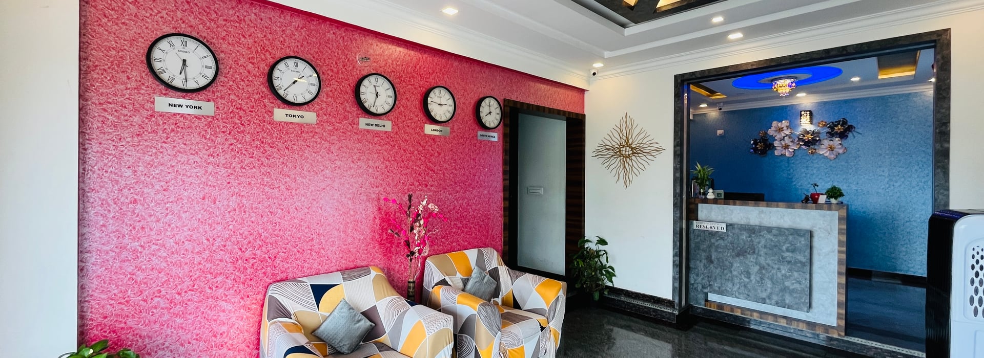 Lobby seating at Value Inn, Bangalore Airport with a patterned sofa, world clocks on a red feature wall, and the reception counter behind