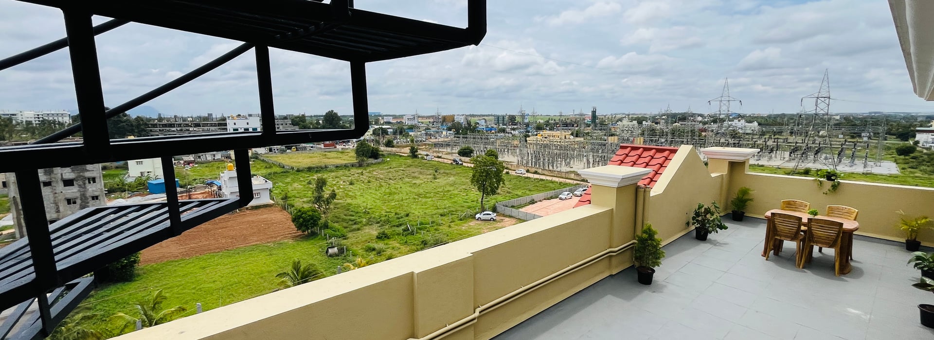 Wide rooftop terrace at Value Inn, Bangalore Airport under a black pergola frame with tiled flooring and open views towards distant fields