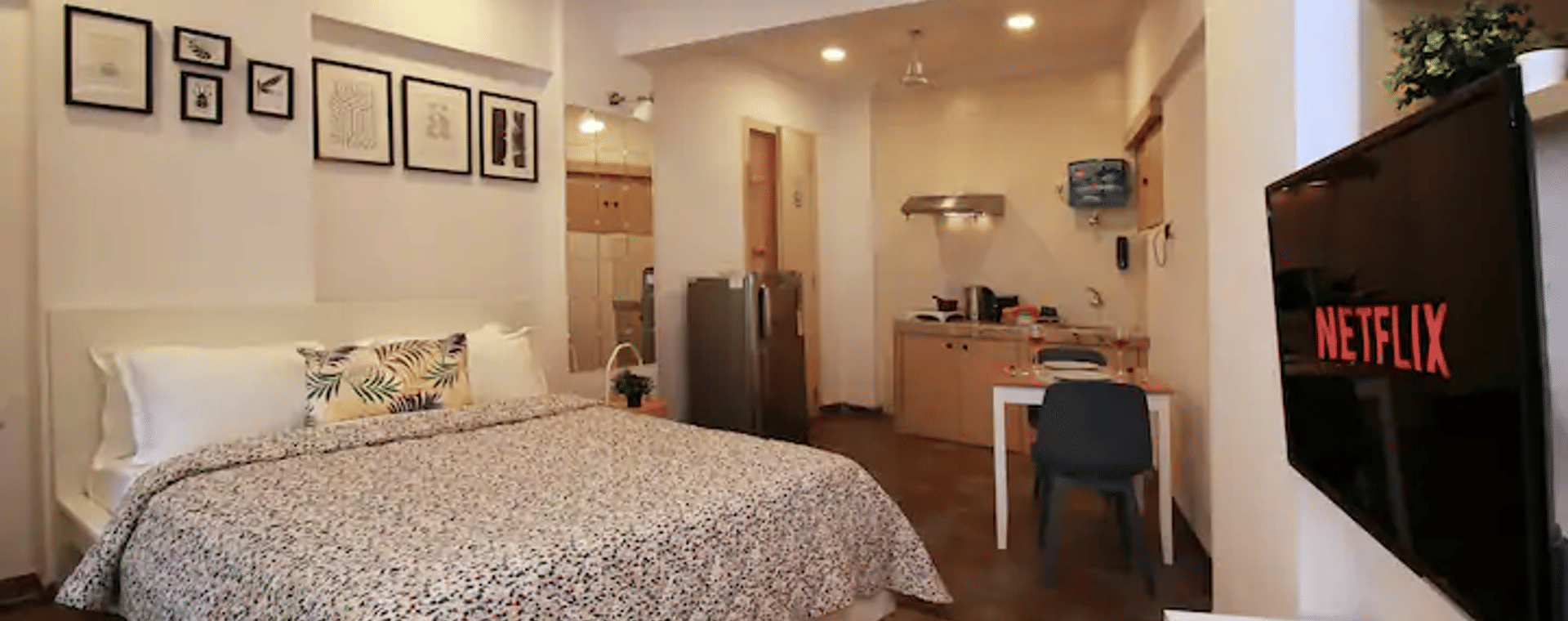 Bedroom of Room 301 at Wabi-Sabi, Juhu, with a printed duvet, wooden flooring, a wall-mounted TV showing the Netflix logo, and an open kitchenette beyond.