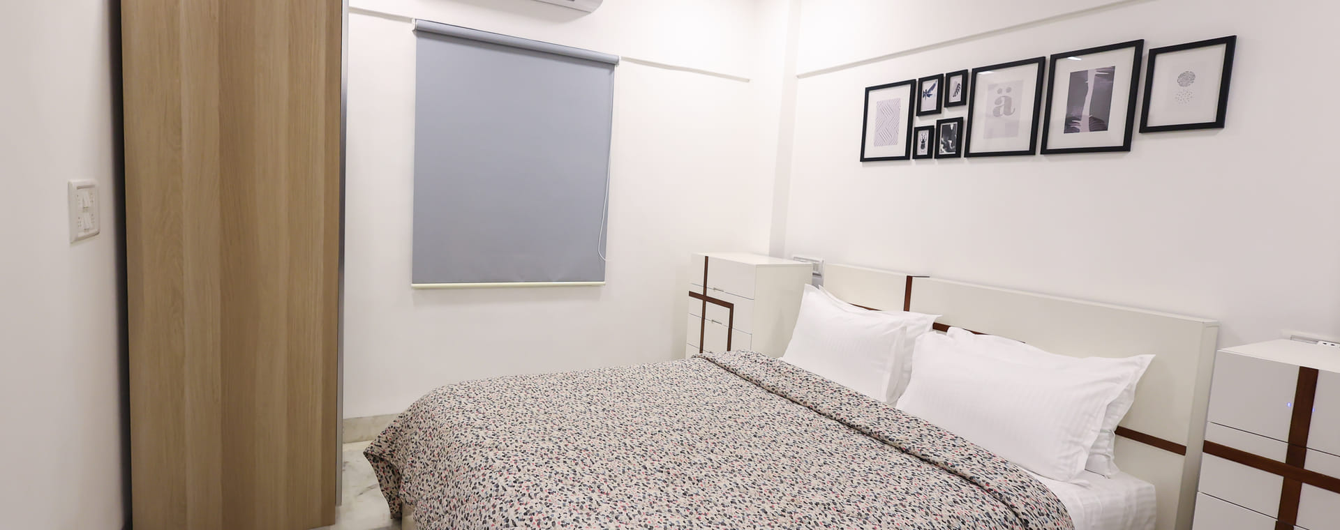 A wide view of the Room 401 bedroom at Wabi-Sabi, Juhu with a patterned bedspread, a split air-conditioning unit, a grey roller blind, and an open wooden wardrobe.
