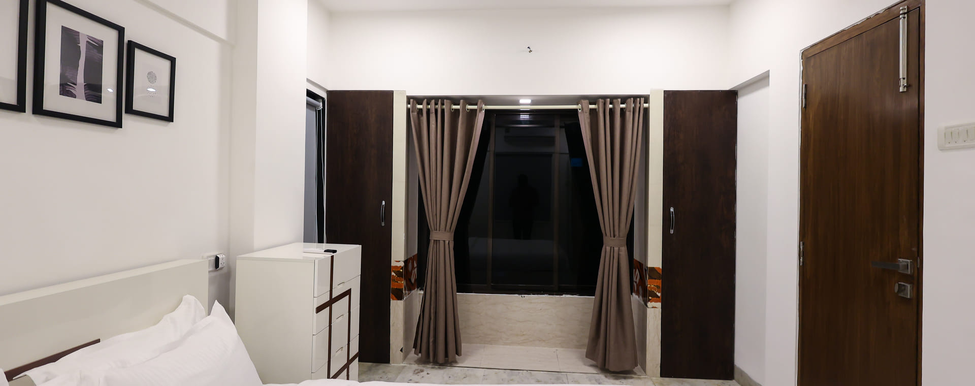 The Room 401 bedroom at Wabi-Sabi, Juhu with a white bed, a wall clock, framed black-and-white photographs, and a curtained window flanked by 2 wardrobes.
