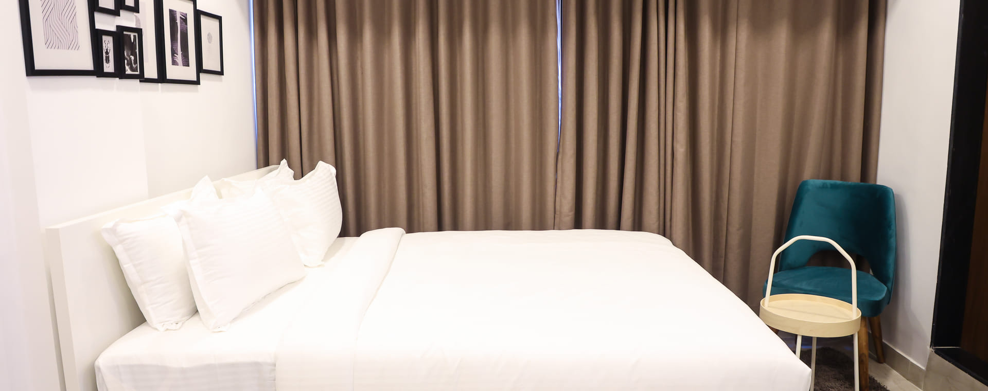 Bedroom of Studio Rooms (E) at Wabi-Sabi, Juhu, with a white bed, brown curtains, a wall-mounted AC, a gallery wall of framed prints, and a teal accent chair.