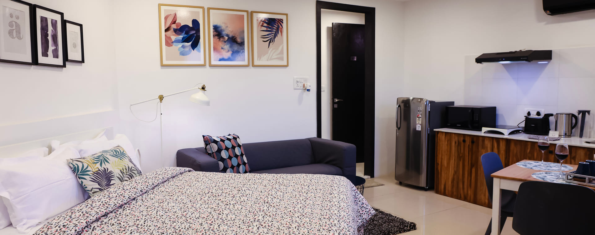 Studio Rooms (B) at Wabi-Sabi, Juhu, with a patterned bedspread, a navy sofa, framed art, and a kitchenette with a fridge, microwave, and dining table.