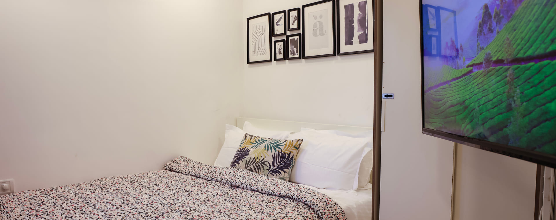 Bedroom of Room 502 at Wabi-Sabi, Juhu, closer view of the leaf-print cushion and headboard beside a wardrobe with an arrow decal and a TV showing tea fields.