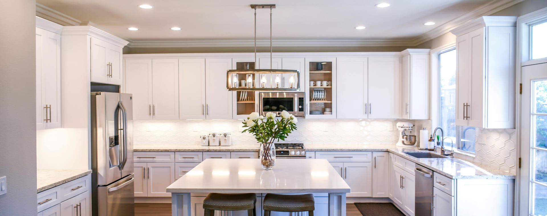 A spacious modular kitchen with white cabinetry, a central island with 2 bar stools, a stainless steel refrigerator, and pendant lighting overhead.