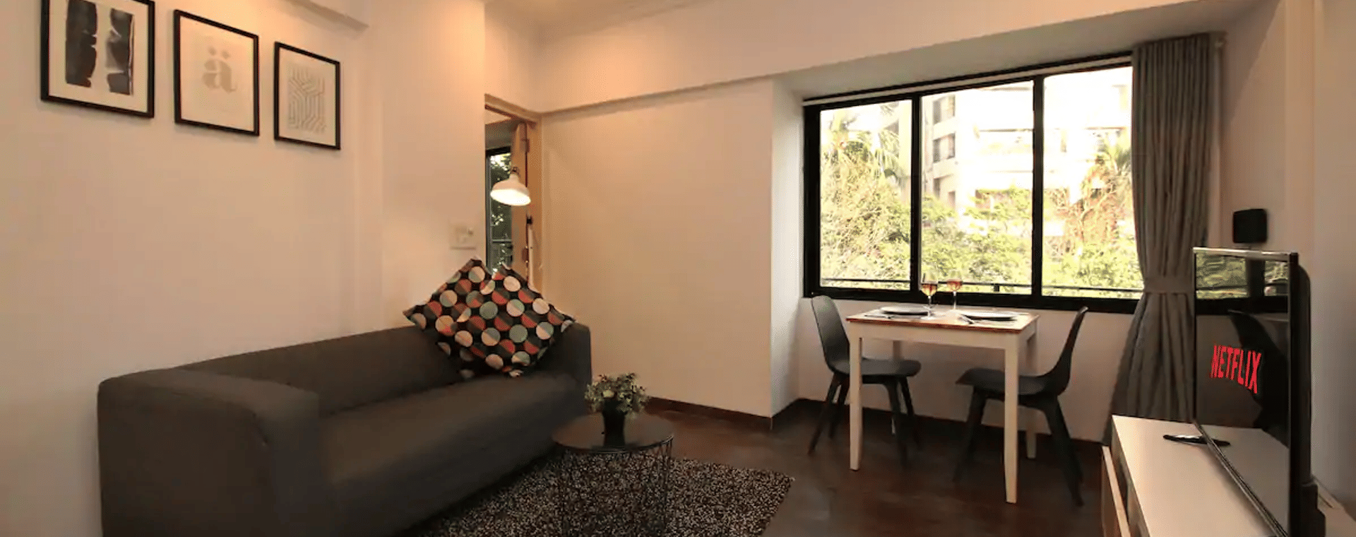 Living room of the 1BHK apartment at Wabi-Sabi, Juhu, with a grey sofa, framed art, a TV console, and a 2-seater dining table beside a window with wine glasses.