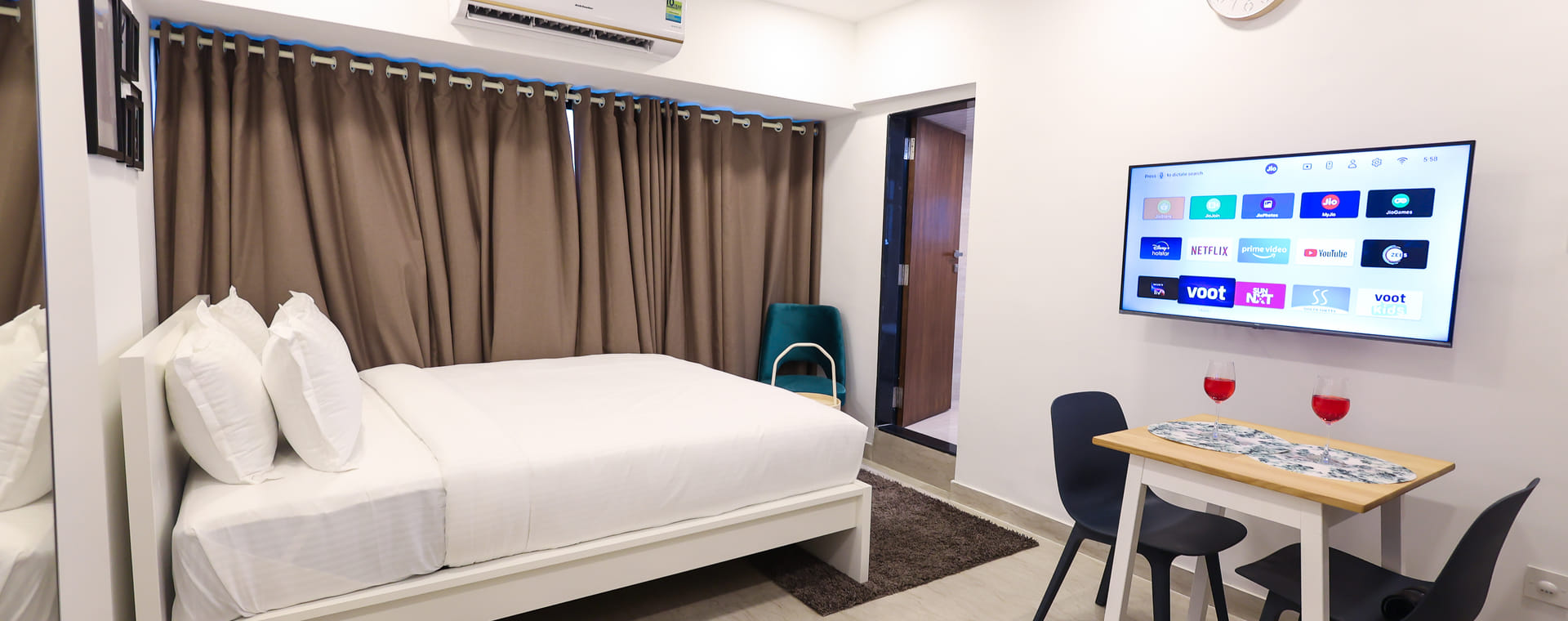 Bedroom at Wabi-Sabi, Juhu, with a bed, brown curtains, a wall-mounted AC, a smart TV showing streaming apps, and a dining table with 2 chairs and wine glasses.