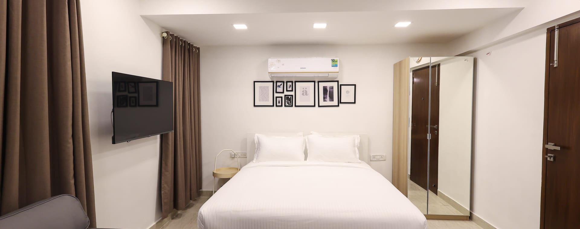 Bedroom at Wabi-Sabi, Juhu, with a white bed, a wall-mounted AC, framed wall art, a mirrored wardrobe, a wall-mounted TV, and a grey sofa.