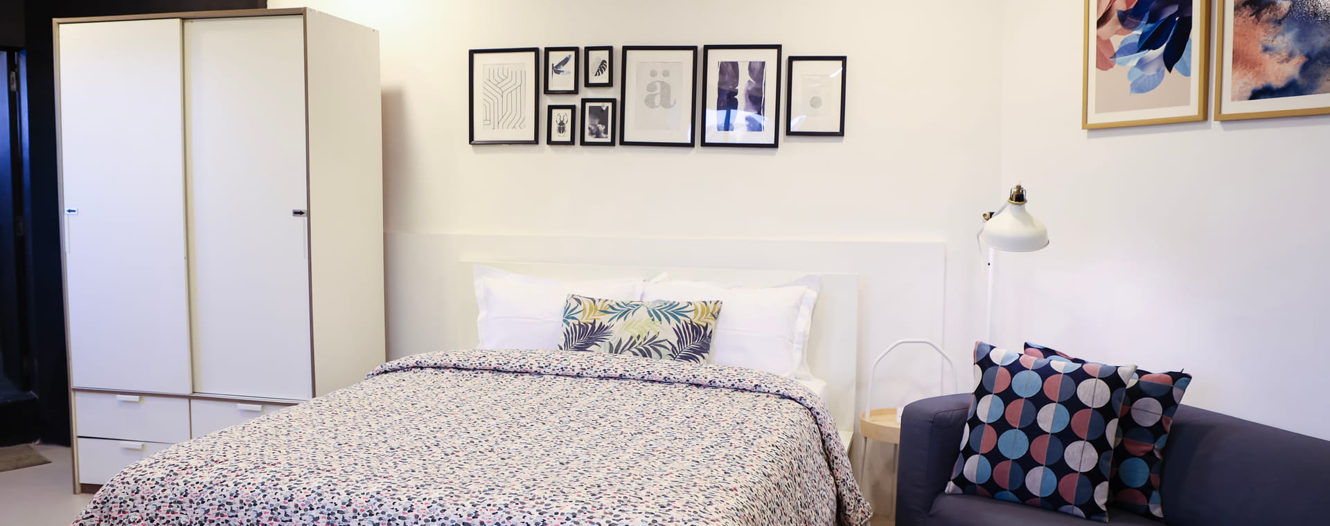 Bedroom at Wabi-Sabi, Juhu, with a patterned bedspread, a white wardrobe, a gallery wall of framed prints, and a navy sofa with printed cushions.