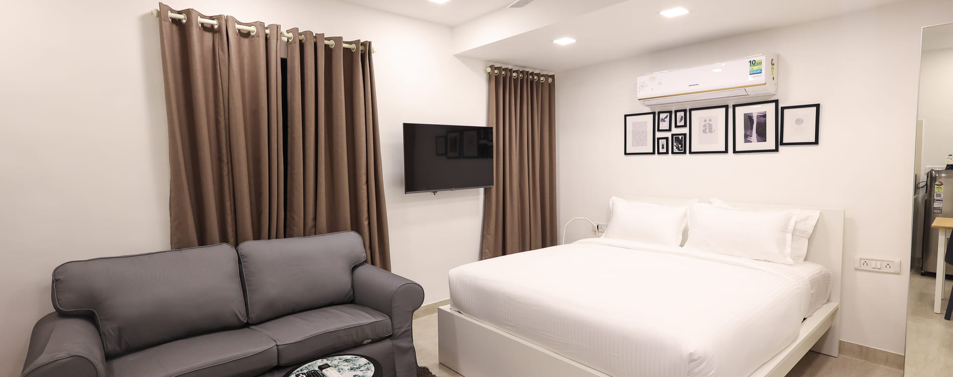 Bedroom at Wabi-Sabi, Juhu, with a white bed, a wall-mounted AC, a gallery wall of framed prints, a wall-mounted TV, and a 2-seater grey sofa.