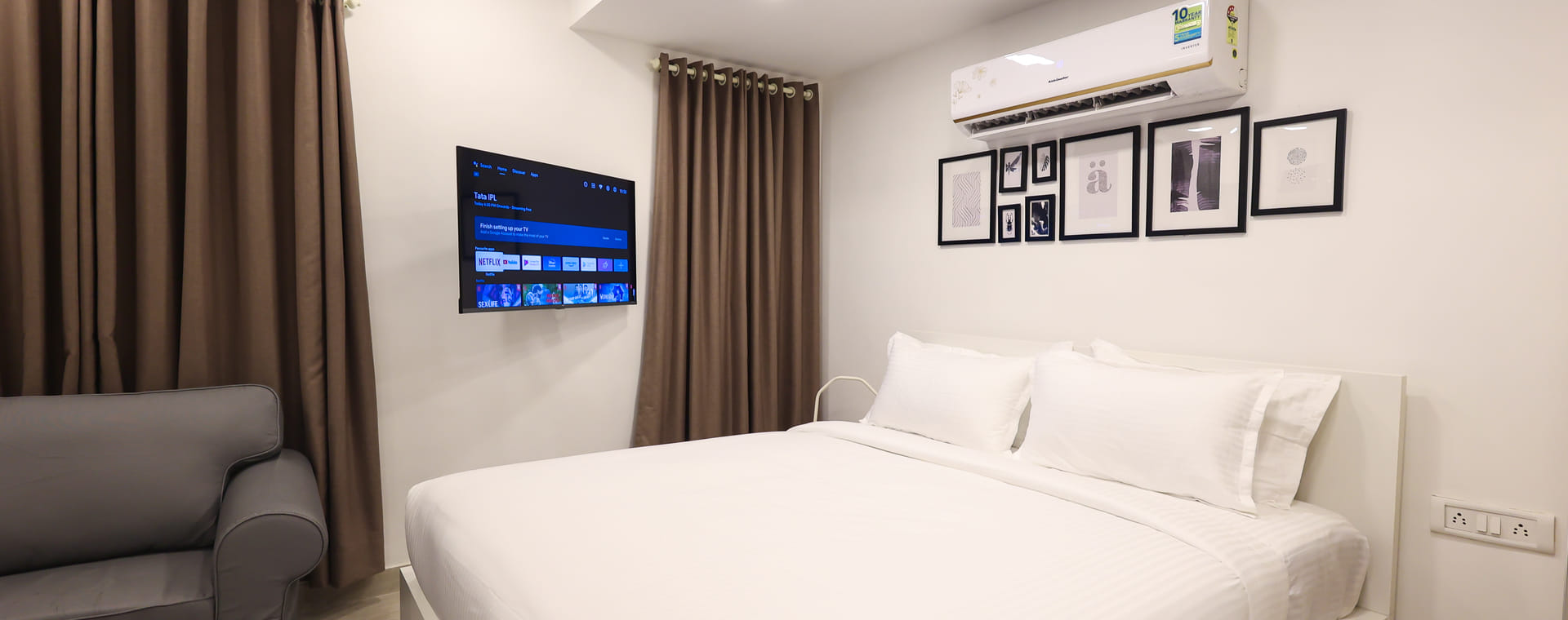 Bedroom at Wabi-Sabi, Juhu, with a grey armchair, brown curtains, a wall-mounted smart TV on its setup screen, and a gallery wall of framed art.