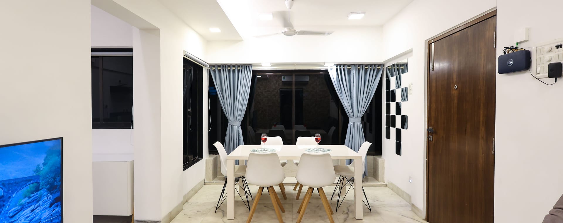 An open dining and living area at Wabi-Sabi, Juhu with a marble floor, a white dining table with 6 chairs, a television on a low console, and a grey sofa.