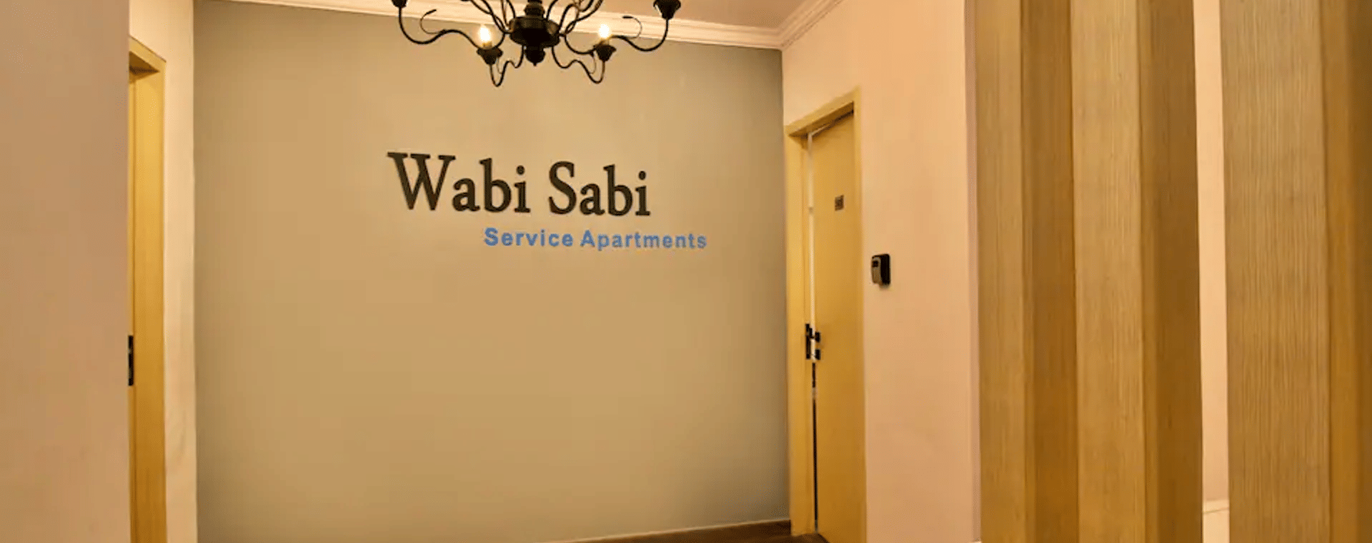 A wooden-floored entrance corridor at Wabi-Sabi, Juhu with a chandelier hanging overhead and a Wabi-Sabi Service Apartments sign on a grey feature wall.
