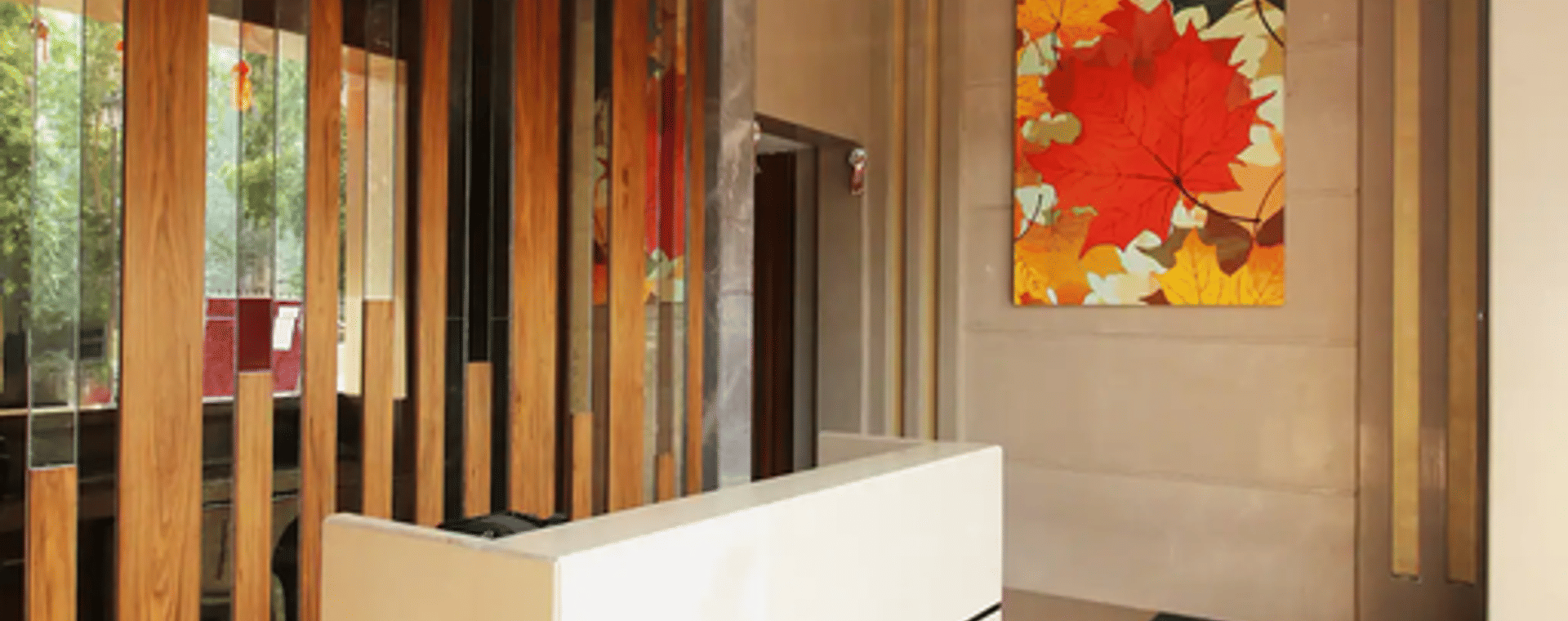 The reception lobby at Wabi-Sabi, Juhu with a white front desk, wooden slat partition, a large autumn-leaf wall art, and a grey carpeted floor.