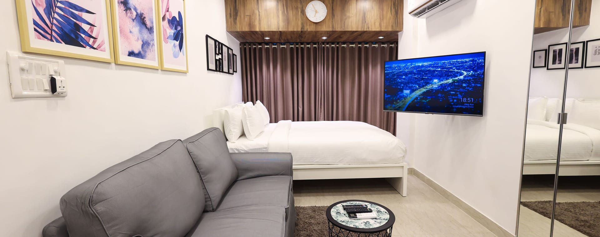 A fully furnished studio apartment at Wabi-Sabi, Juhu with a grey sofa, white bed, wall-mounted television, mirrored wardrobe, and a wooden headboard with a clock.