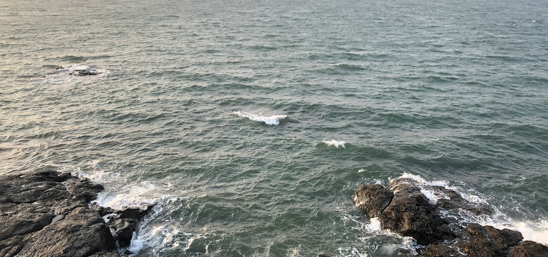 Waves breaking over dark rocks along the Malvan shore with a hazy headland across the water | Shirgaonkar Beach Resort