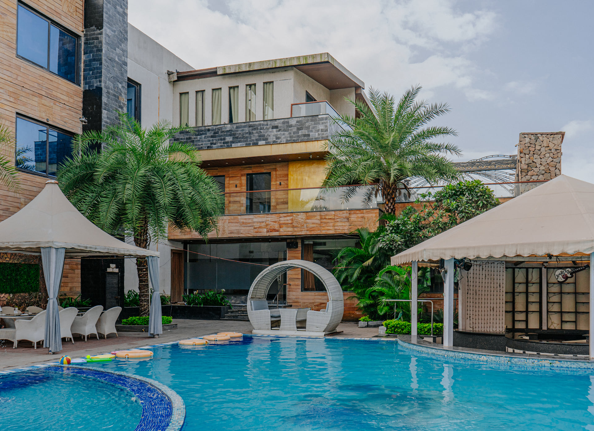 Swimming pool in front of the stone-clad hotel building with palm trees and white gazebos