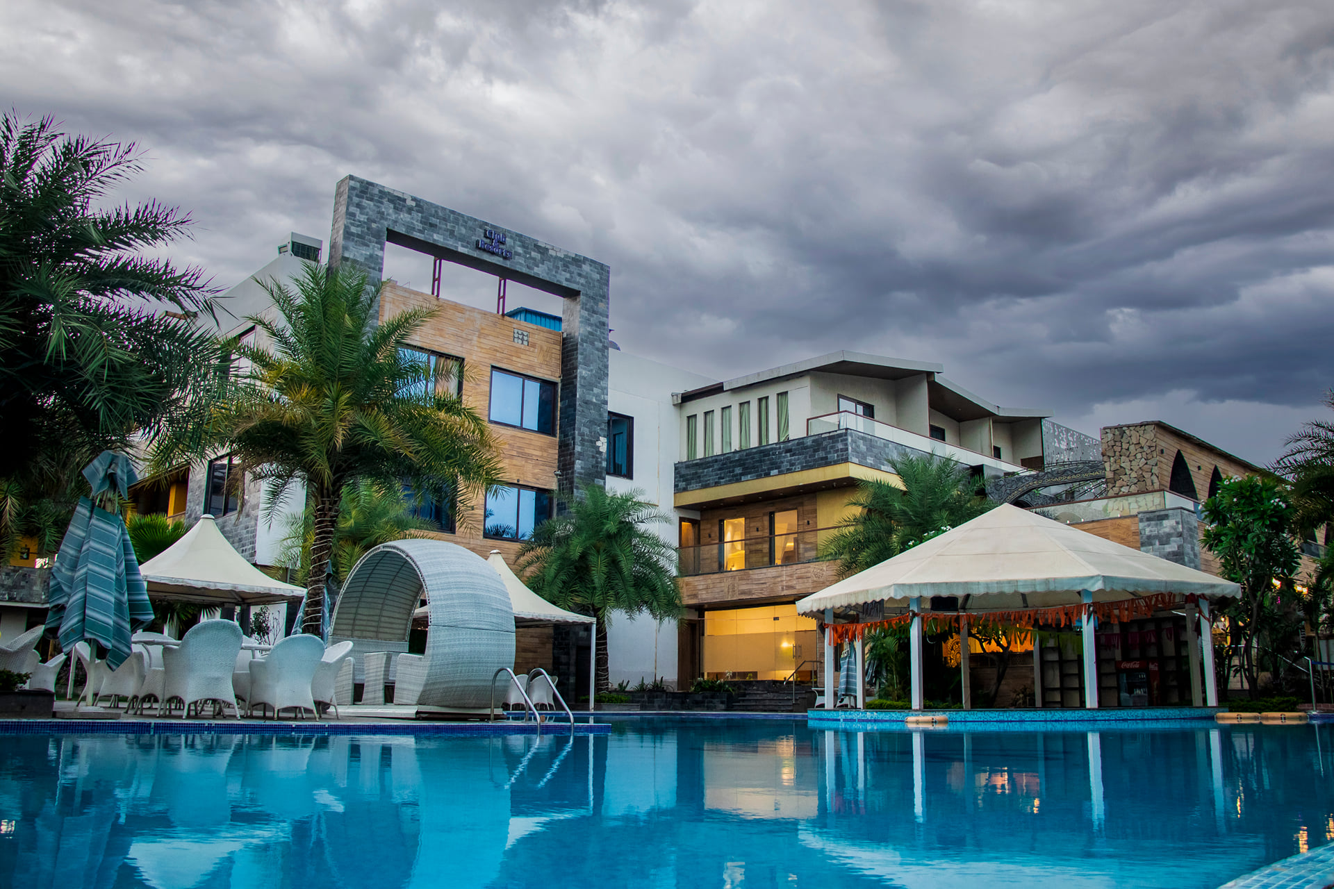 Hotel buildings and a white poolside canopy beyond the swimming pool under a cloudy sky, with palm trees