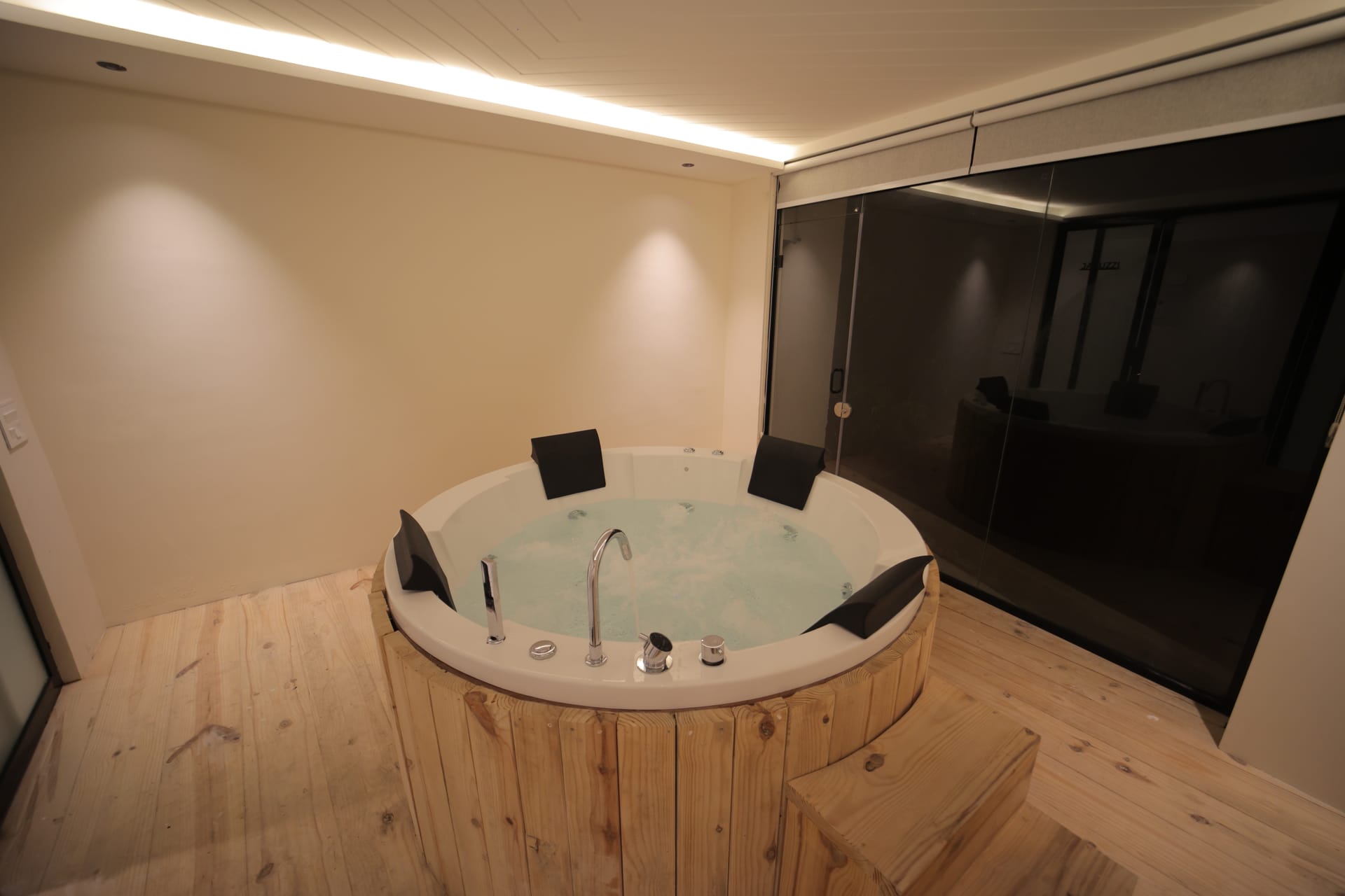 A wooden-framed Jacuzzi tub at Nazaara Hills Retreat, Udaipur, lit softly at night against a mirrored glass wall.