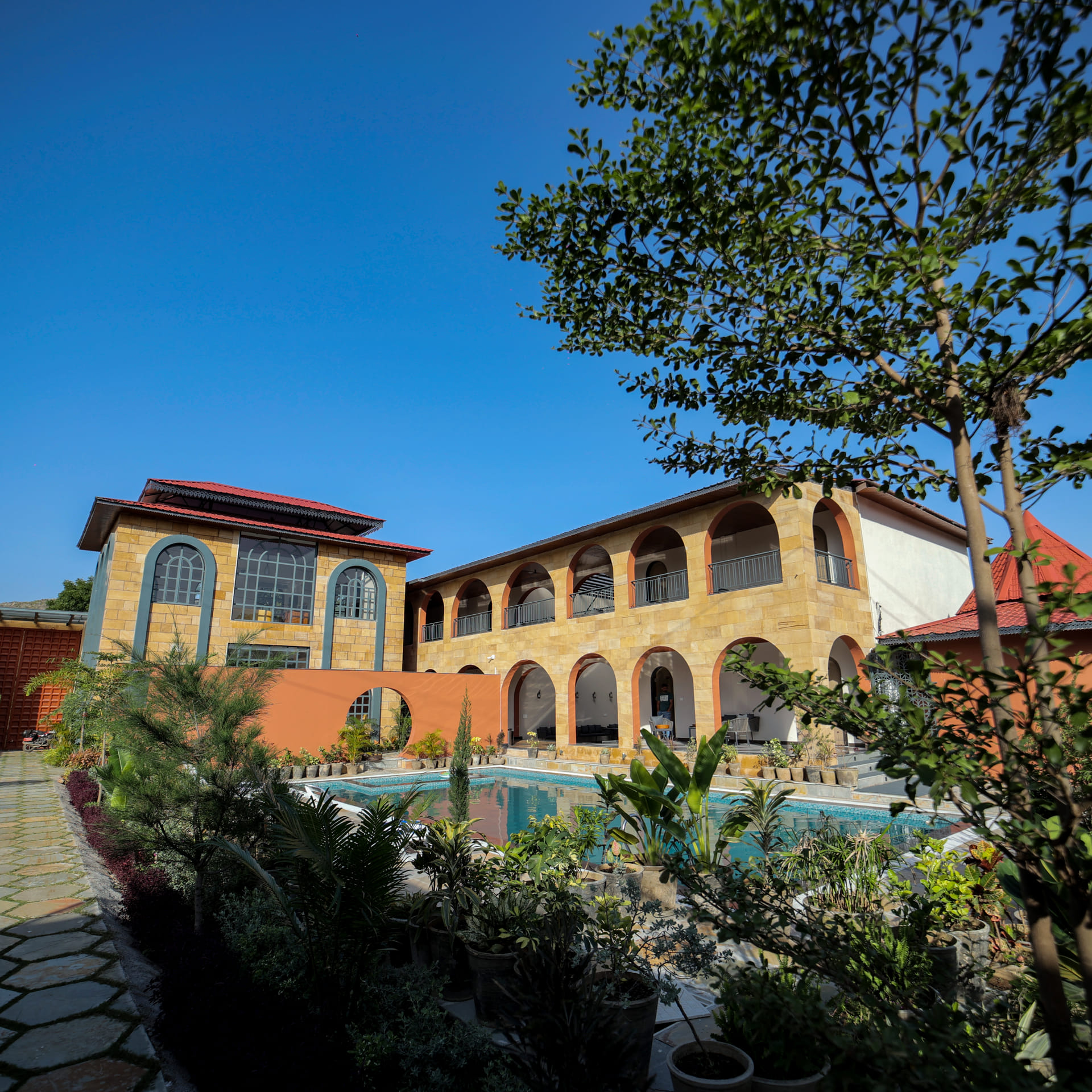Arched sandstone facade and courtyard pool at Nazaara Hills Retreat, Udaipur, framed by garden greenery under a clear sky
