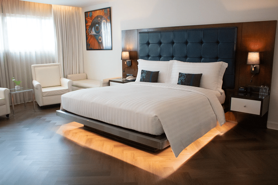 An overview of the Grand Gold King Room's bedroom with illuminated platform bed, tufted headboard, vibrant artwork, cream armchairs, and warm ambient lighting throughout at Night Hotel Bangkok in Sukhumvit - the best district to stay in Bangkok.