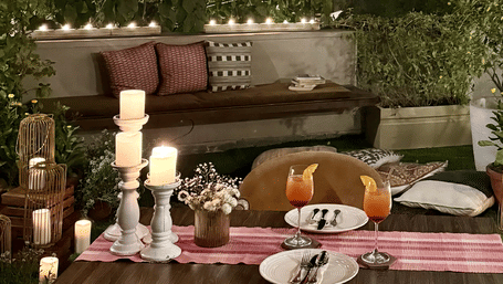 A dinner setting at the outdoors featuring cold drinks, cutlery and plates, flower vase, and lit candles on table and a seating area with cushions.