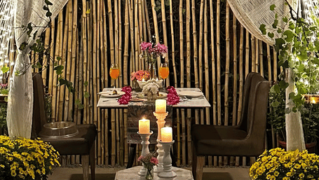 A romantic outdoor dining setup at Karma Chalets with a draped canopy, bamboo backdrop, candles, flowers, and warm lights creating an intimate evening ambience amidst lush greenery.