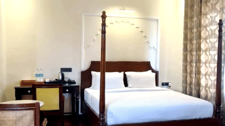 A room with a large bed, spreads, pillows, a fan, and a table. | Hotel Devnadi, Haridwar