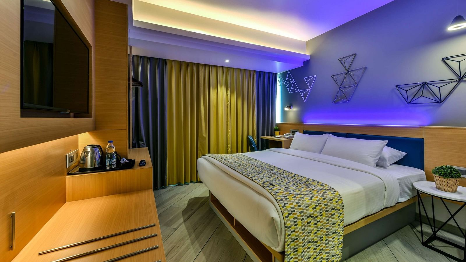 A spacious room with a large bed, a wall-mounted television, a desk positioned below it, and windows with curtains drawn, illuminated by cool lighting at Indie Stays, BKC Kalina.