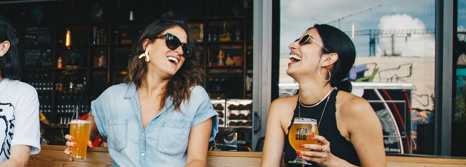 Two woman enjoying holding a glass of cold beverage as they laugh while sitting on a wooden bench.