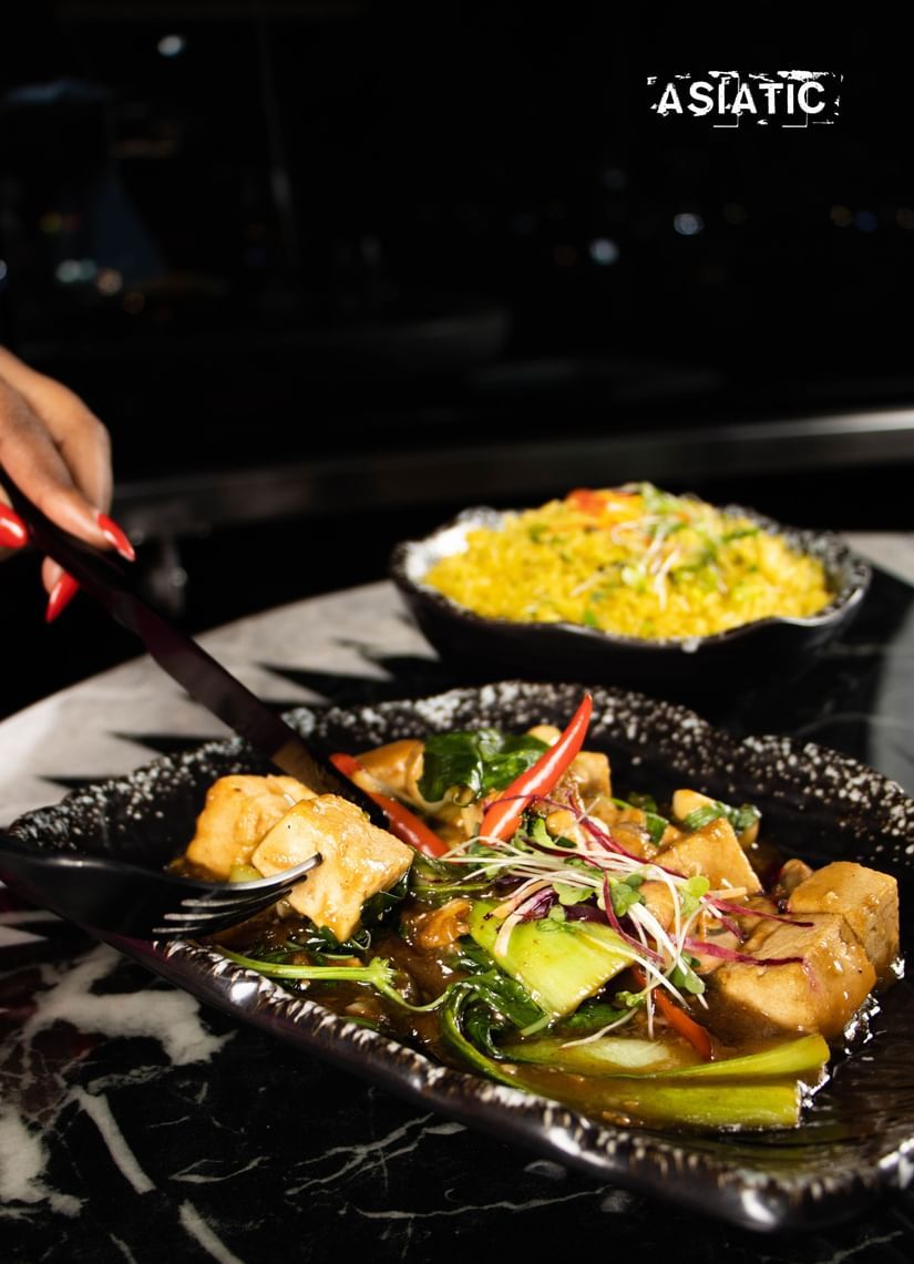 Plate with food items being served, with a hand holding a spoon at Asiatic Lounge.