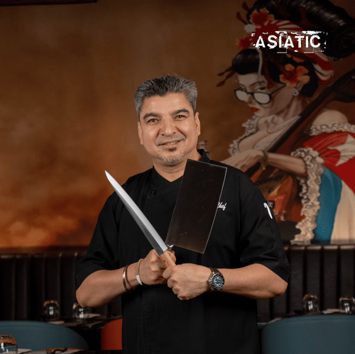 Chef stands holding knives in front of restaurant seating at Asiatic Rooftop Bar and Restaurant.