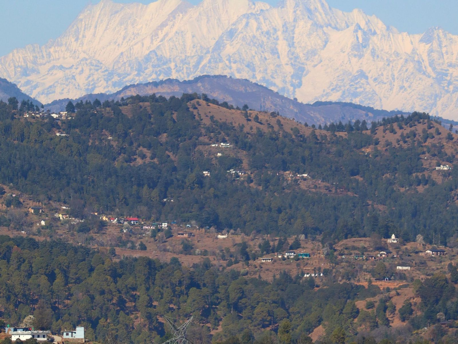 A clear view of snow-capped mountains with green valleys and buildings visible in the foreground under a bright blue sky.