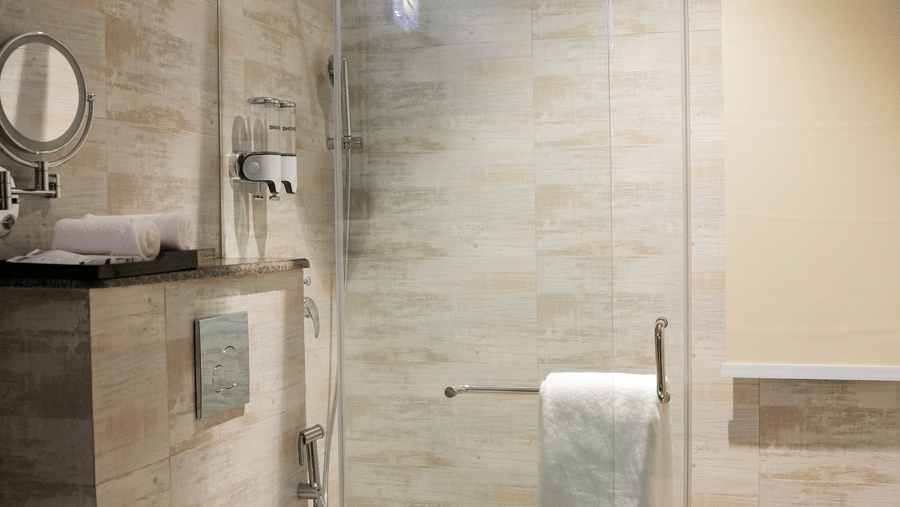 A well-lit bathroom at The Meriton, showcasing a glass-enclosed shower cubical, a commode, a basin, and a ceiling fan, with light-coloured tiled walls.