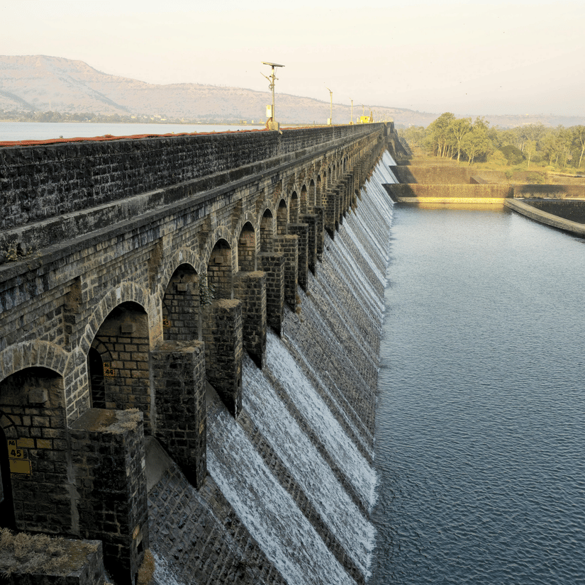 Long dam stretching across a reservoir at sunrise, with calm waters reflecting soft light and distant hills beyond