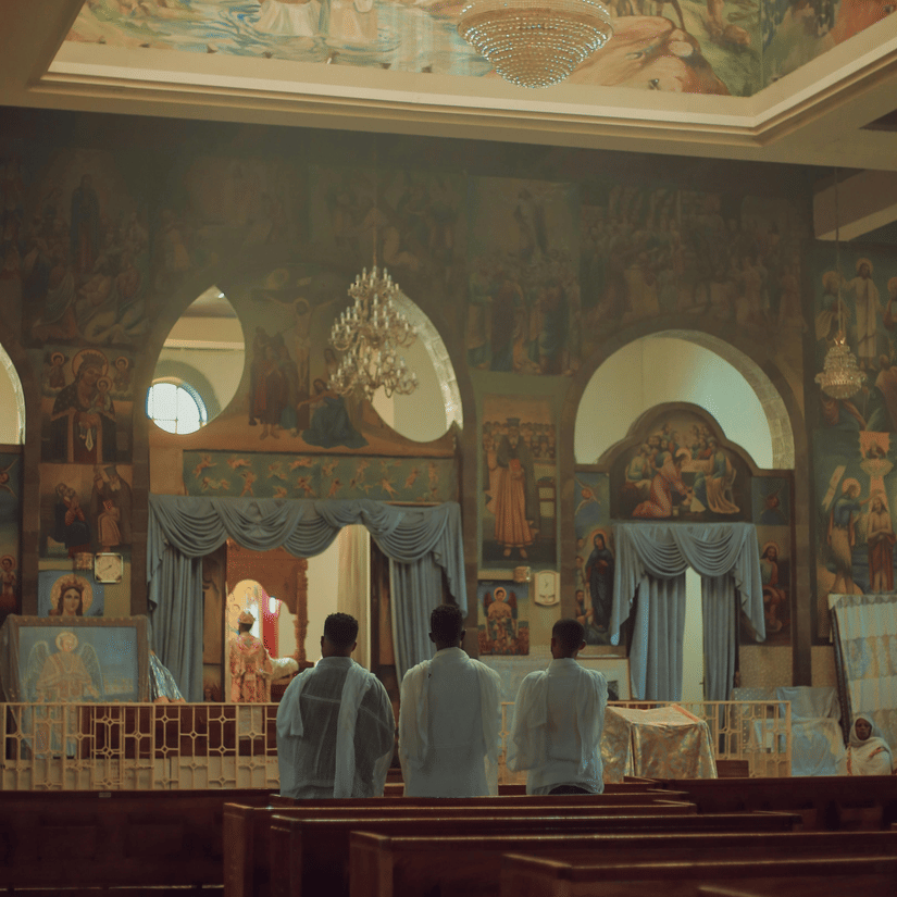 3 people stand in pews facing a wall with arched openings beneath a large, colourful religious ceiling mural.