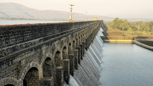 A dam enclosure viewed in the early evening hours releasing small streams of ater from a few gates, with a claar blue sky in the background.