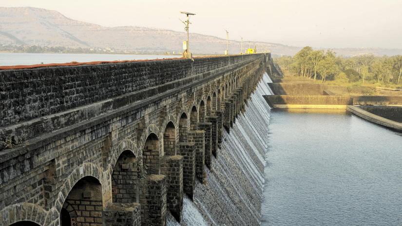 A dam enclosure viewed in the early evening hours releasing small streams of ater from a few gates, with a claar blue sky in the background.