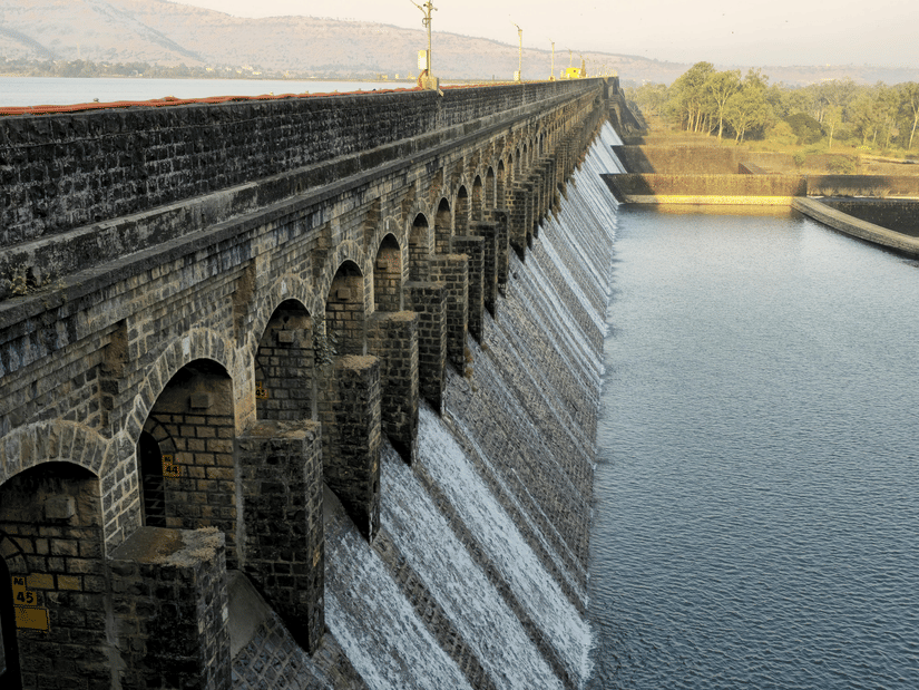 A long stone dam structure with water cascading down its side, next to a calm reservoir under a bright sky.
