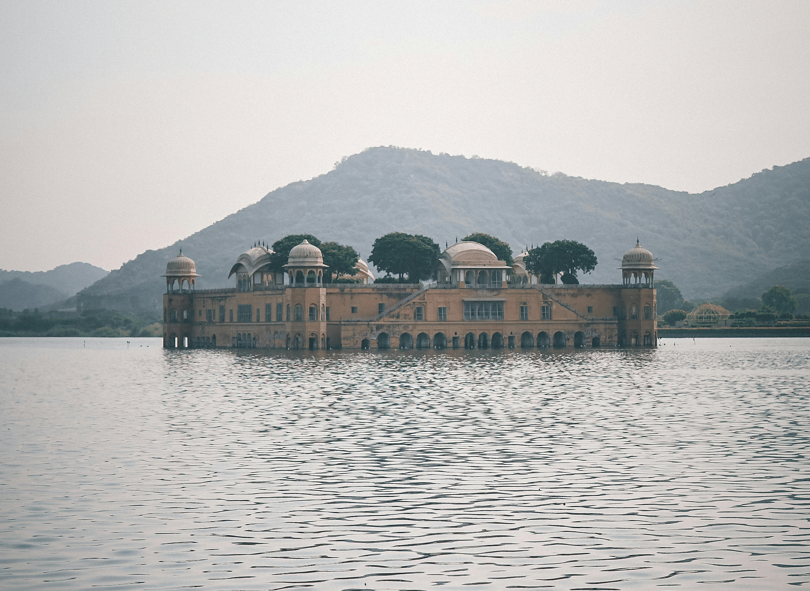 A distant view of Jal Mahal, a palace partially submerged in Man Sagar Lake, is seen under a hazy sky, with a mountain range visible in the background.