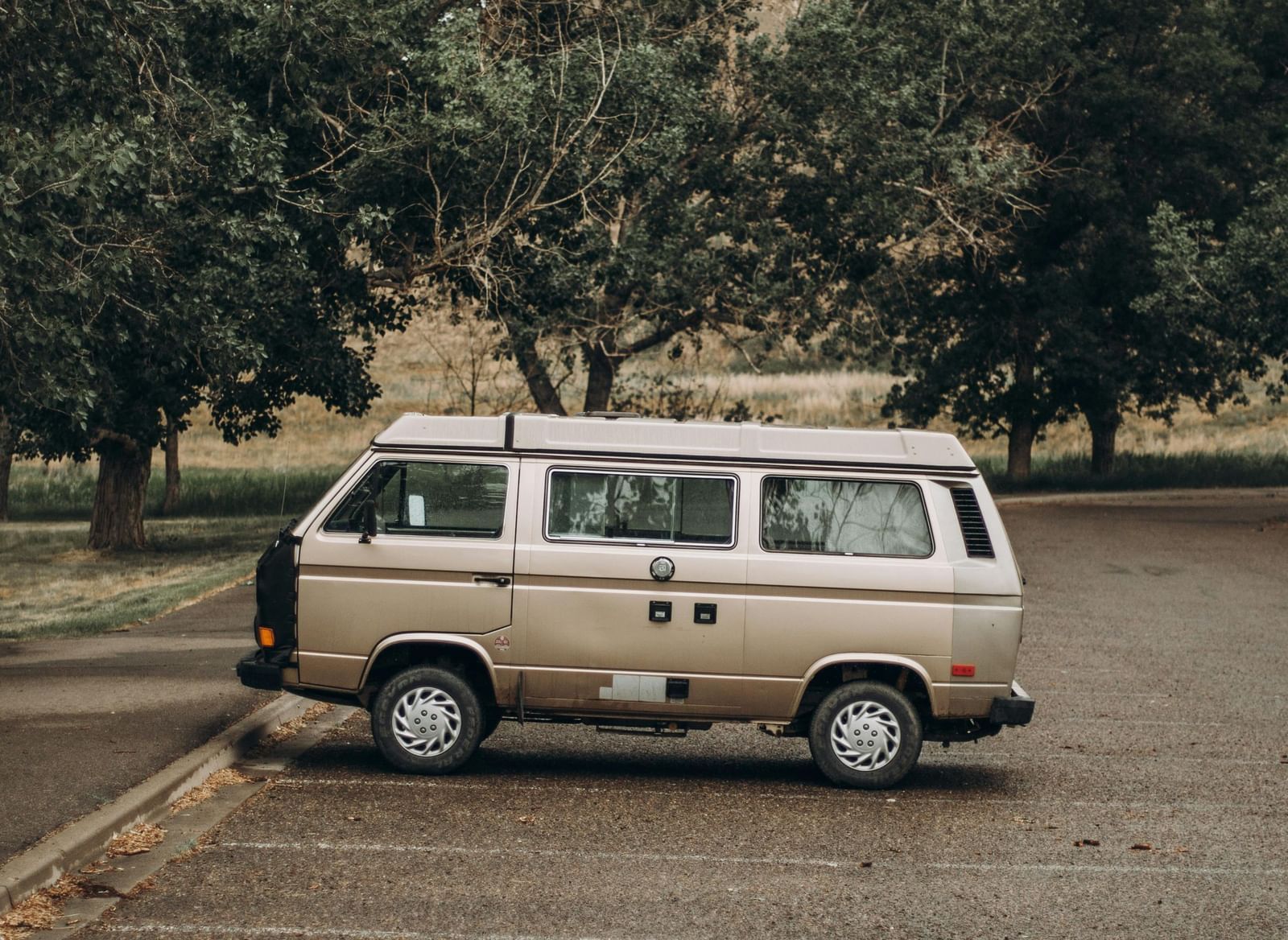 A vintage, light-coloured van parked on a dirt or gravel path, with tall trees and a building visible in the background under an overcast sky.