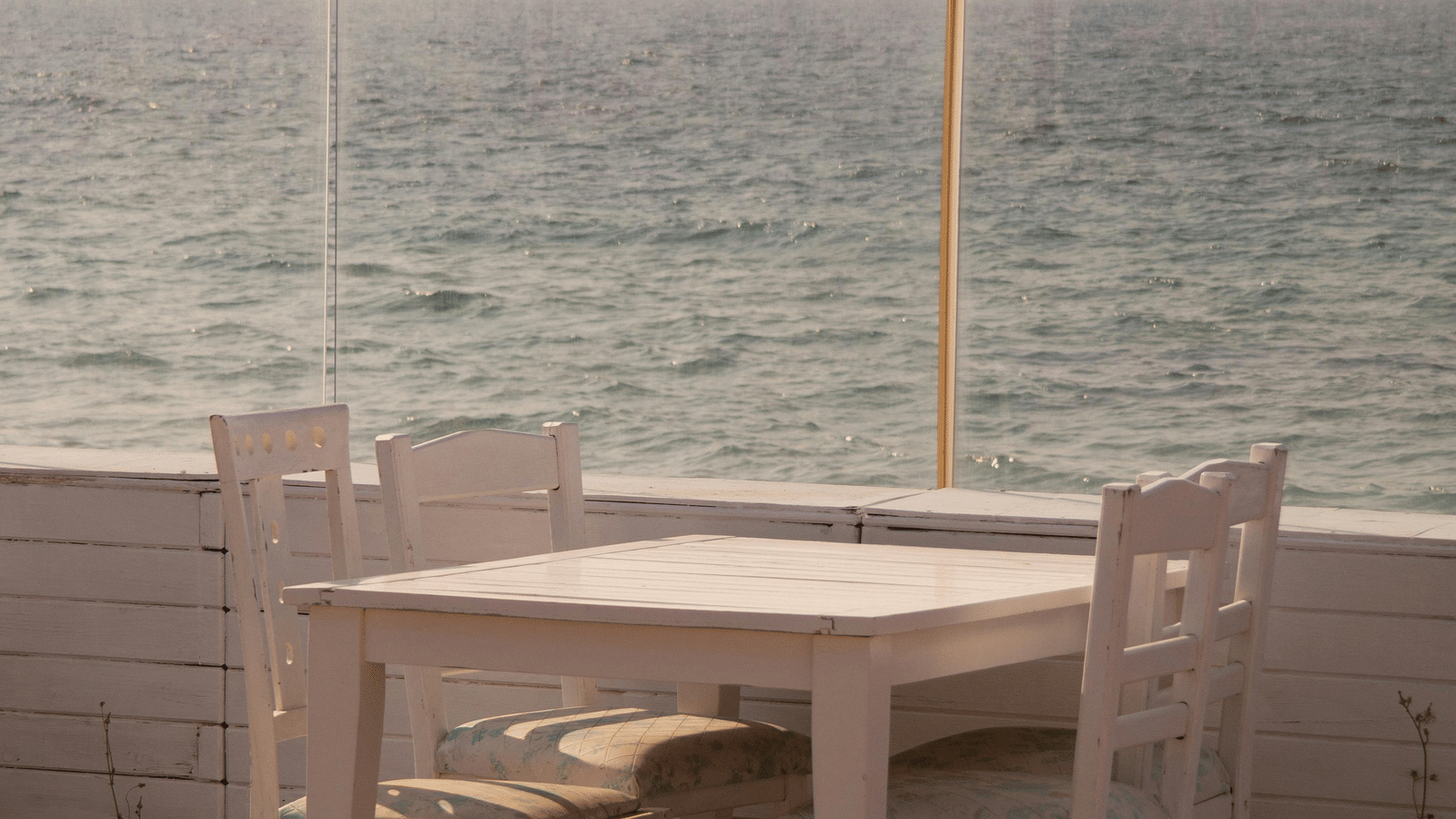 A wooden patio featuring the white table and two chairs set next to a railing overlooking the sea.