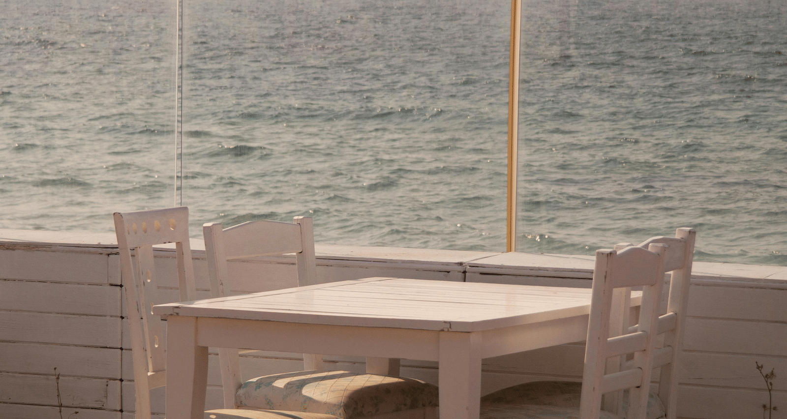 A wooden patio featuring the white table and two chairs set next to a railing overlooking the sea.