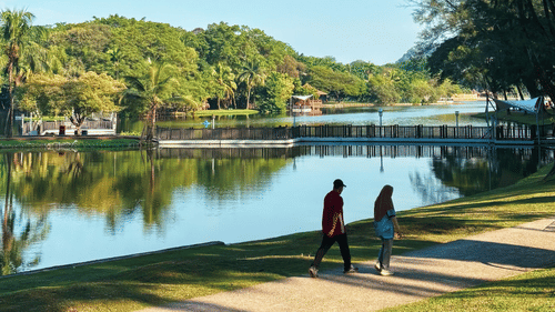 A paved pathway running alongside a calm river features tall green trees, a grassy lawn, and people walking under a bright sky.